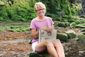 Carol sits on the beach with her sketchbook