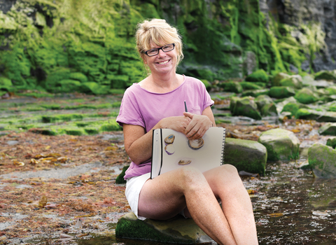 Carol sits on the beach with her sketchbook